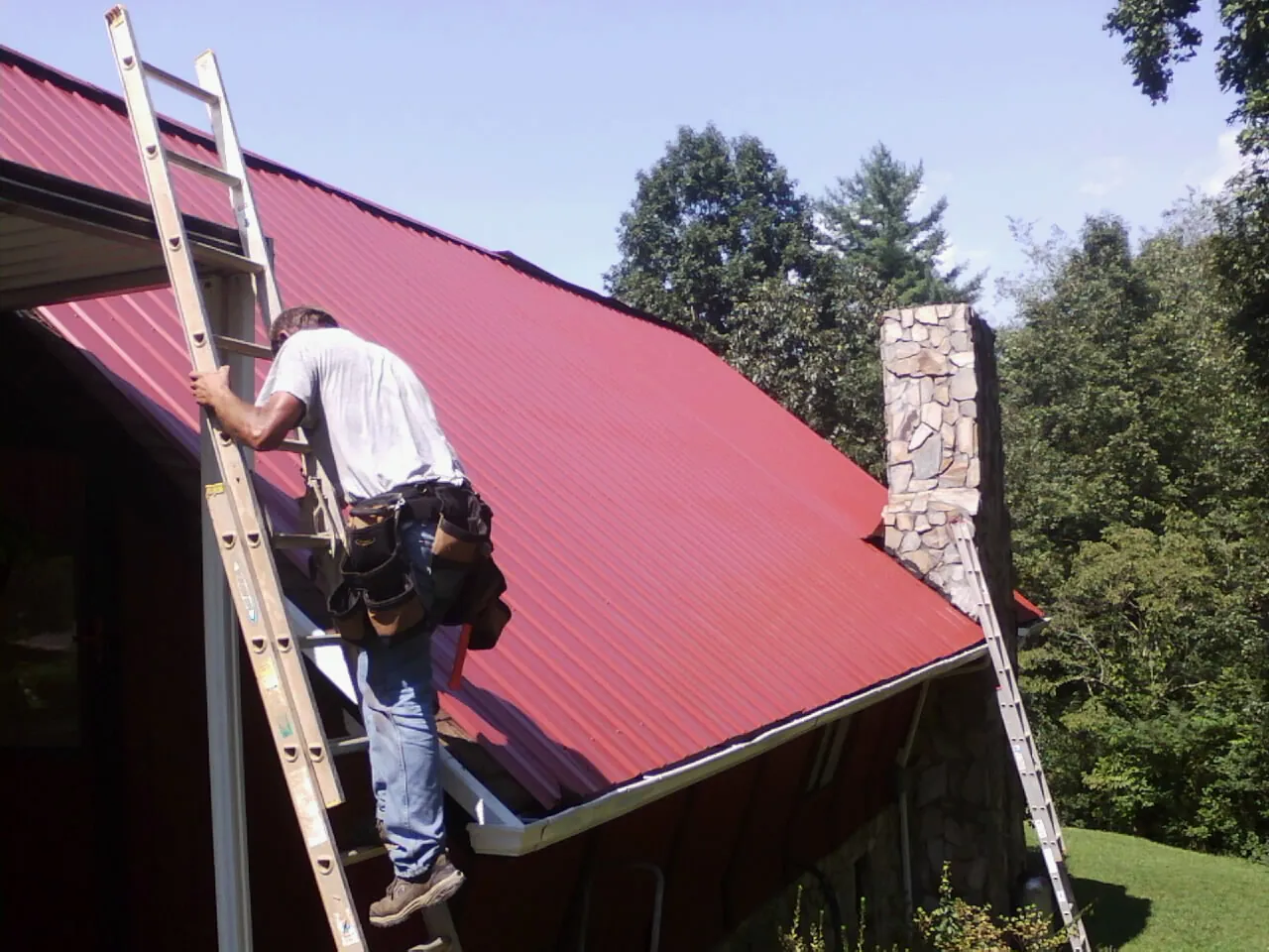Roofer on a ladder beside a red metal roof and stone chimney — Soffit & Fascia Repair by Roofing Brothers in Okolona